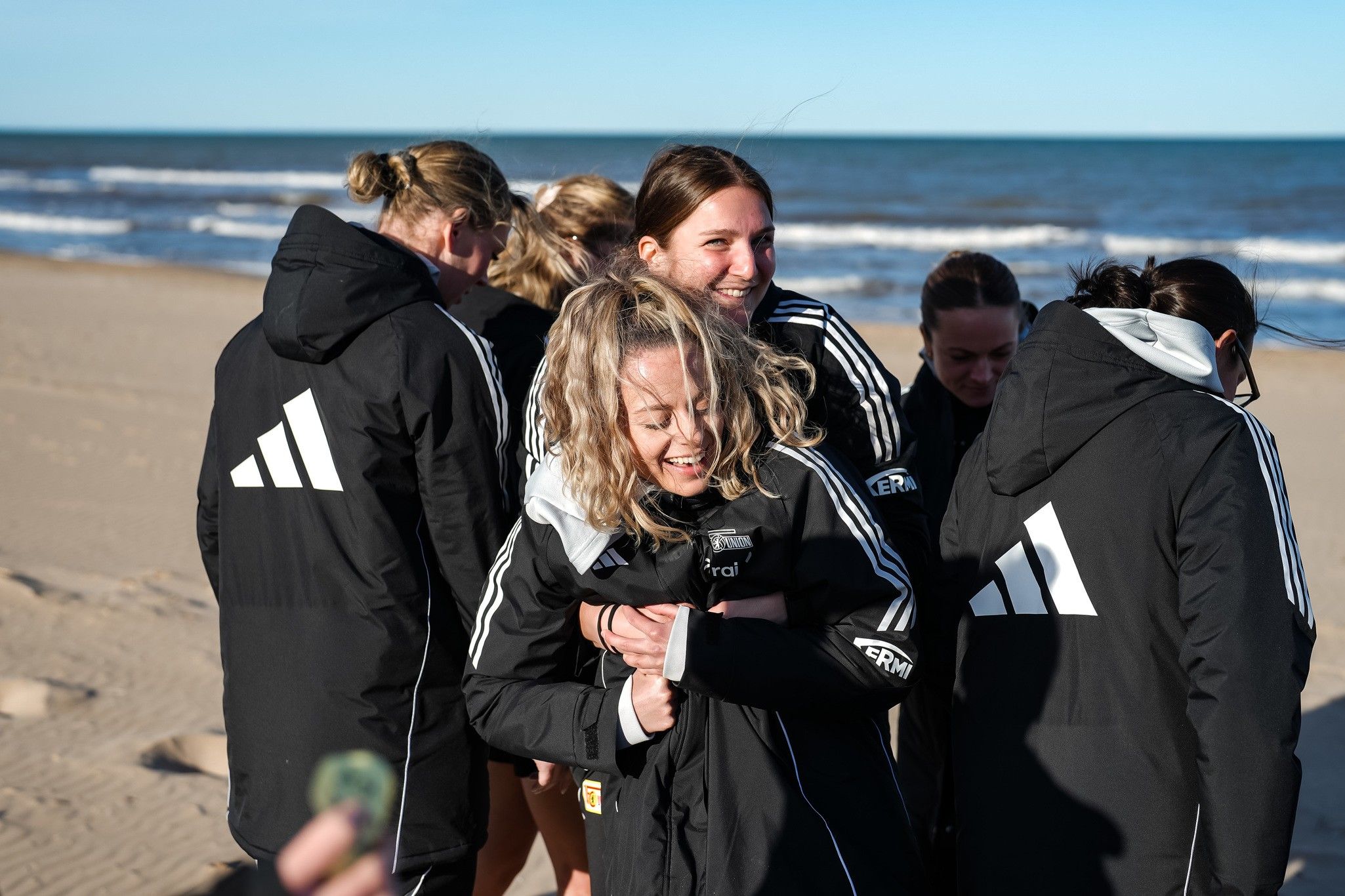 Gruppe von Frauen in sportlichen Jacken am Strand, die lachen und sich umarmen. Meer und Himmel im Hintergrund.