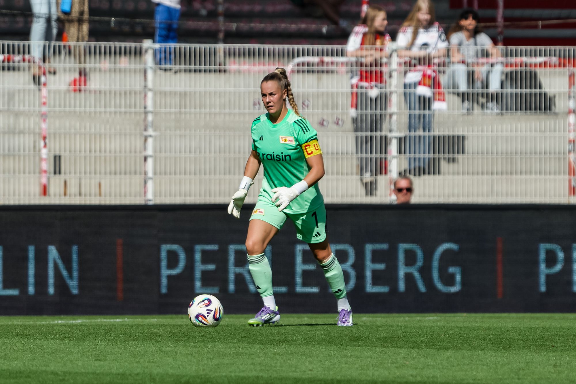 Torhüterin in grünem Trikot mit Kapitänsbinde beim Spiel, in Aktion auf dem Feld. Zuschauer im Hintergrund.