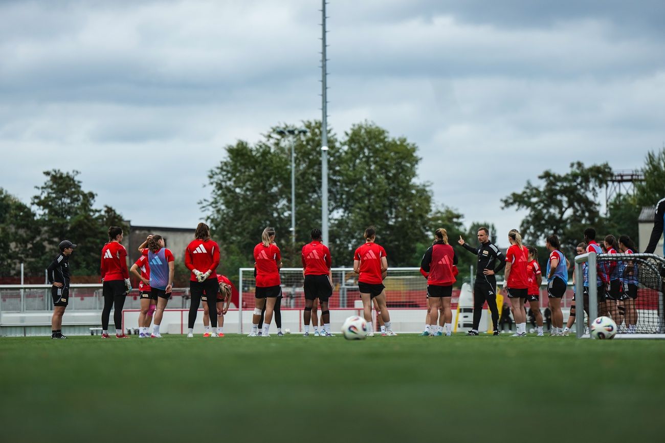 Gruppentraining einer Frauensportmannschaft auf einem Fußballplatz, mit Spielern in roten Trikots und Trainer im Gespräch.