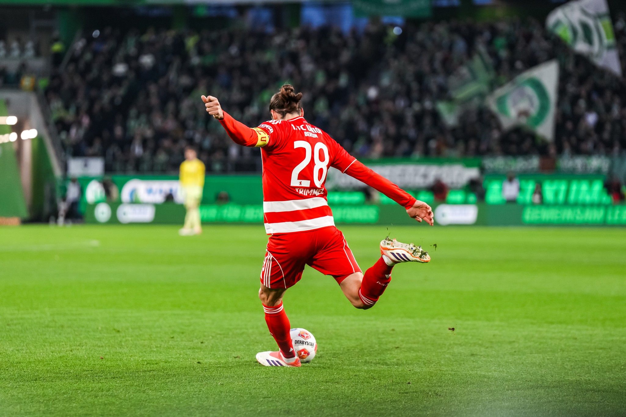 Ein Fußballspieler in rotem Trikot mit der Nummer 28 tritt den Ball auf einem Spielfeld vor einer jubelnden Menge.