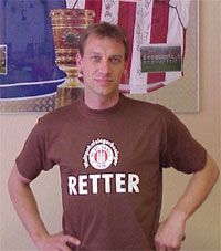 Mann in braunem T-Shirt mit der Aufschrift "RETTER" steht vor einer Wand mit Fußballtrophäen und -trikots.
