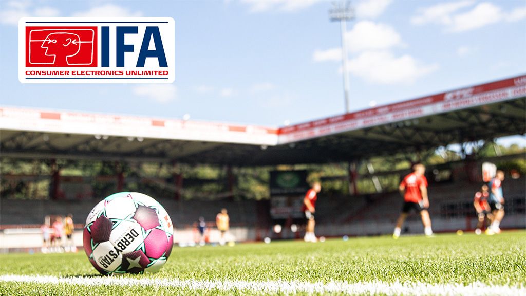 Ein Fußball liegt auf einem grünen Rasenfeld, während im Hintergrund Spieler trainieren und ein Stadion sichtbar ist.