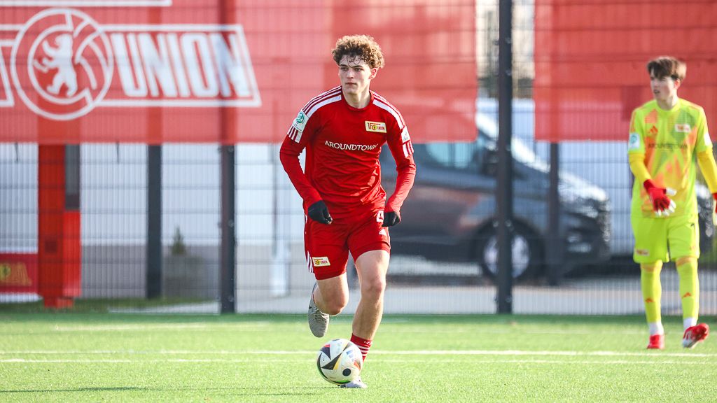 Ein junger Fußballspieler in rotem Trikot läuft über ein Kunstrasenfeld, während ein Torwart im Hintergrund steht.