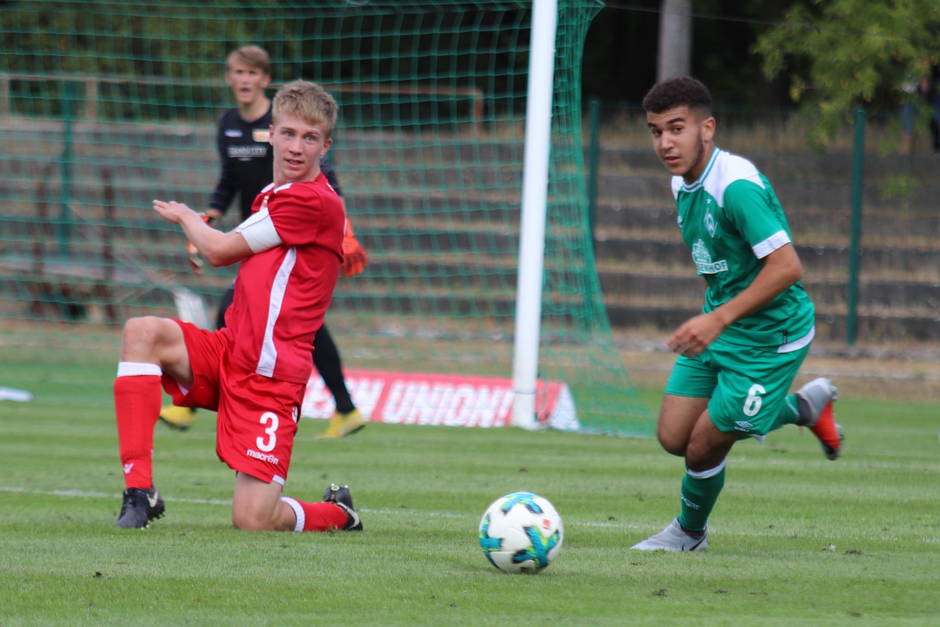 Zwei Fußballspieler im Wettkampf: ein Spieler in roter und einer in grüner Uniform, im Hintergrund ein Tor und Spielfeld.