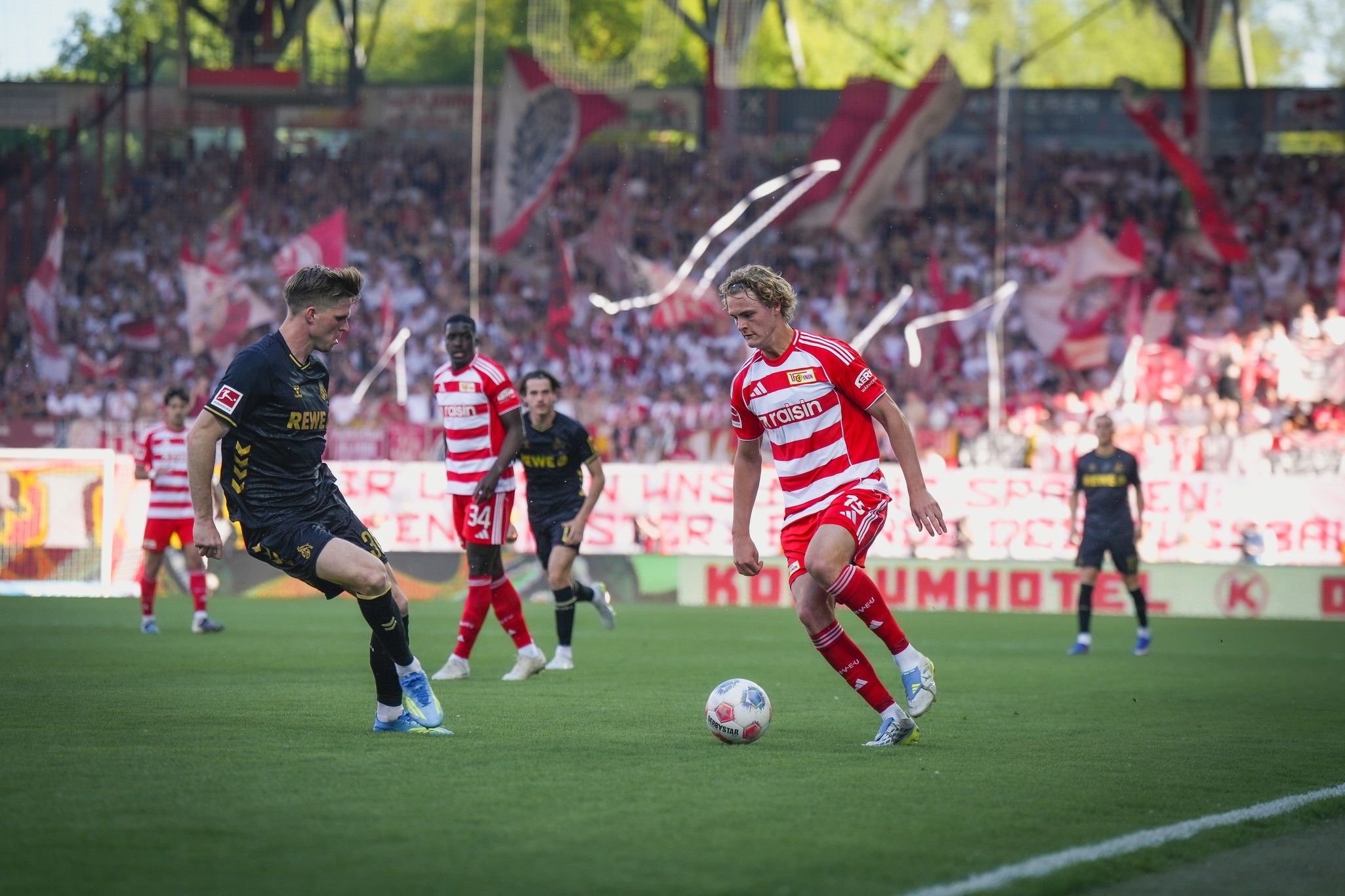 Spielszene aus einem Fußballspiel mit zwei Spielern, die den Ball auf einem Rasenfeld verfolgen, umgeben von jubelnden Fans.