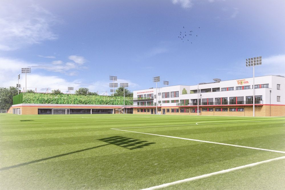 Sportplatz mit Rasen, Trainingsgebäude im Hintergrund und Hochlichtmasten, unter blauem Himmel.