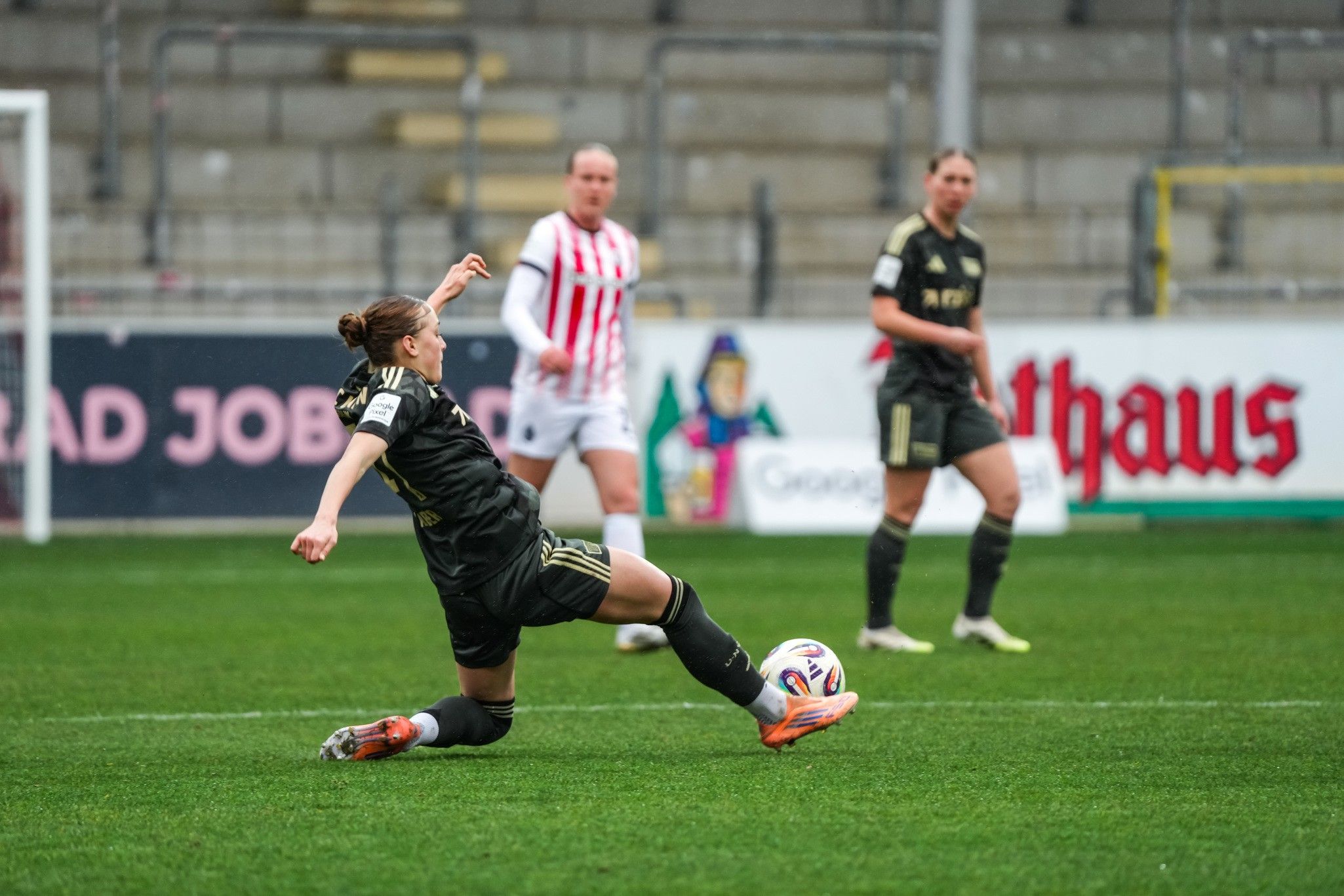 Eine Fußballspielerin rutscht im Rasen, während zwei andere Spielerinnen im Hintergrund stehen.