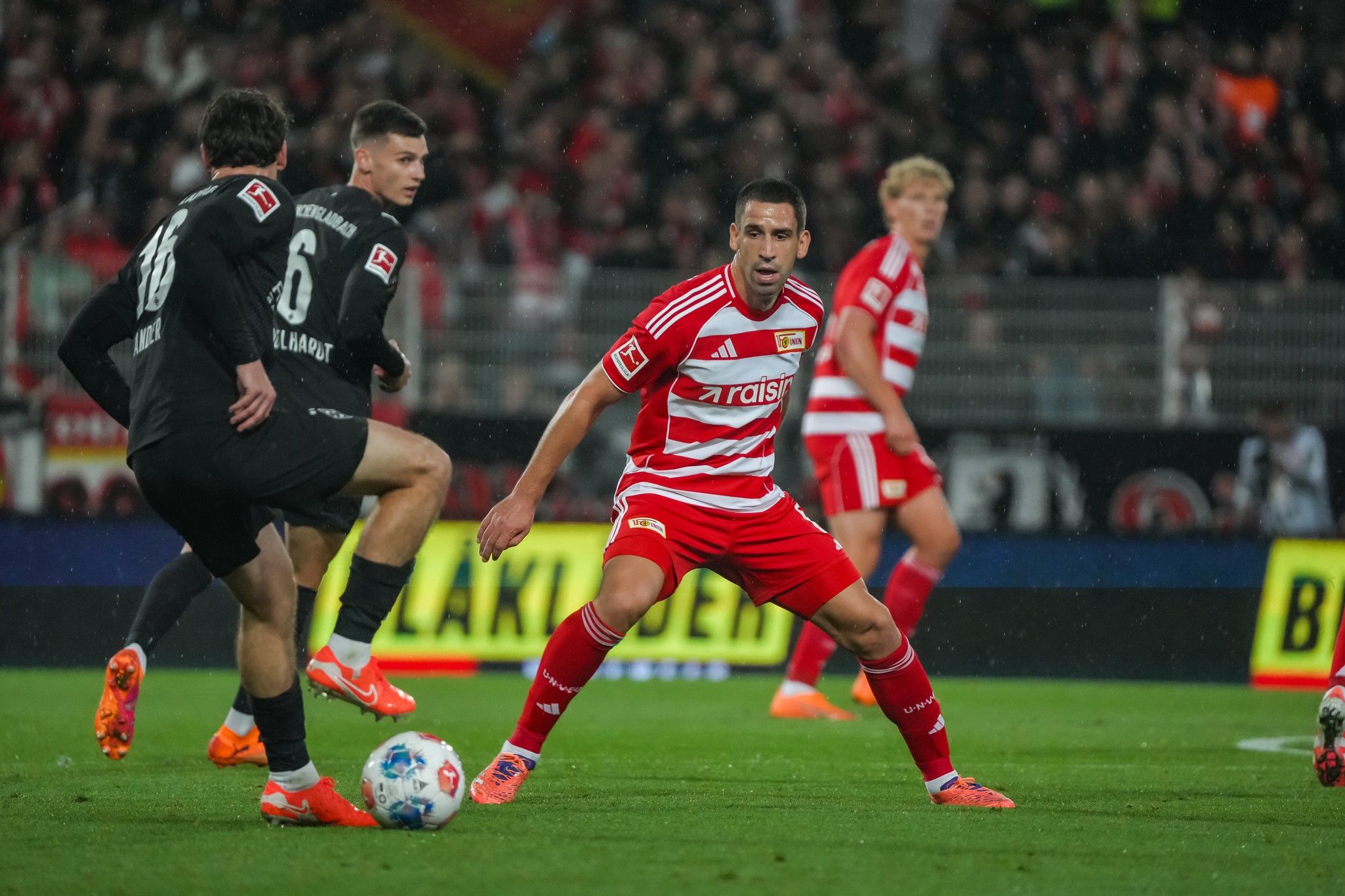 Ein Fußballspieler in roter und weißer Uniform am Ball, während ein Verteidiger in Schwarz ihm entgegenkommt. Stadionhintergrund.