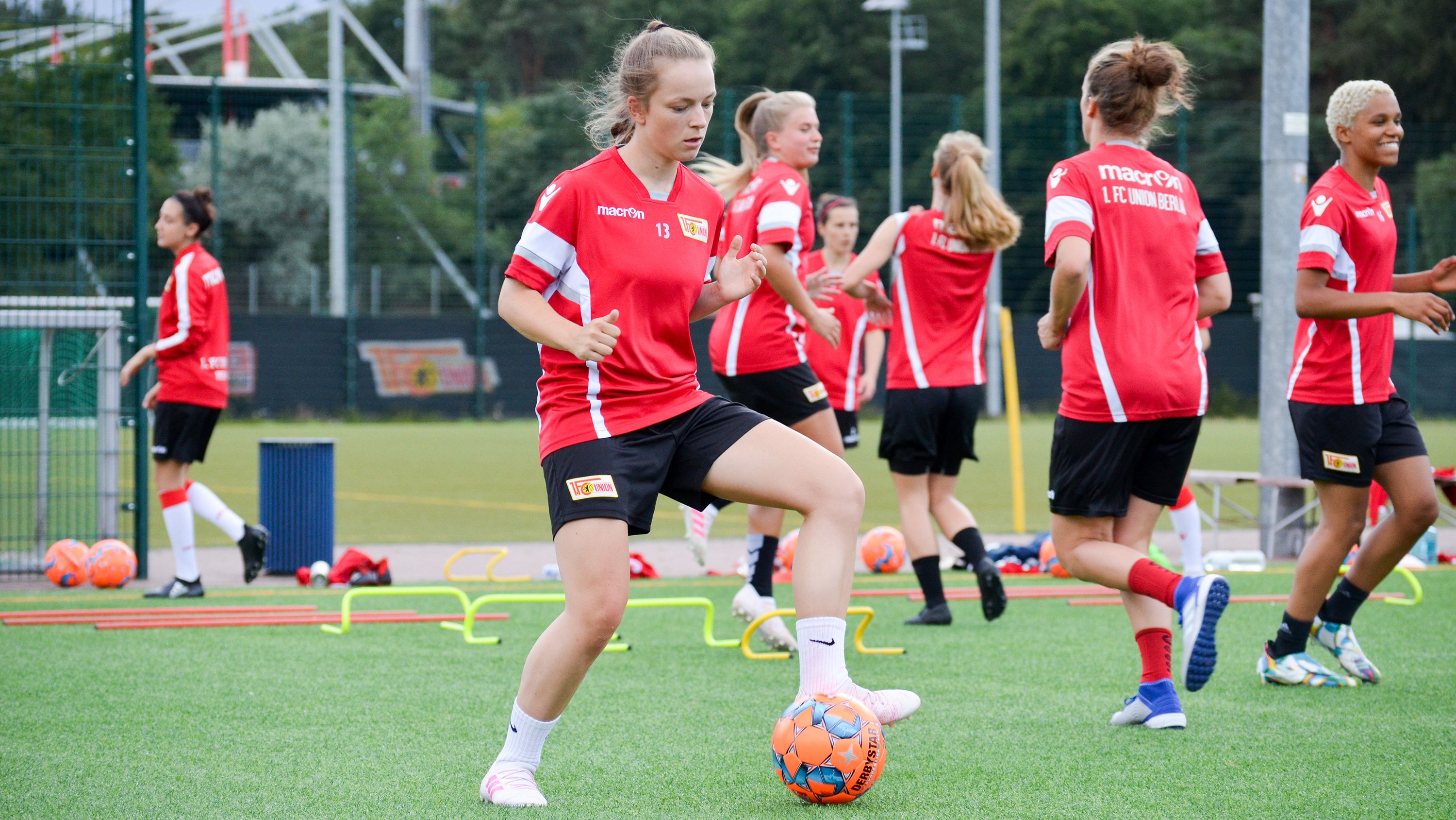 Mädchen in rotem Trikot trainieren Fußball auf einem Kunstrasenplatz mit orangen Bällen und Trainingsutensilien.