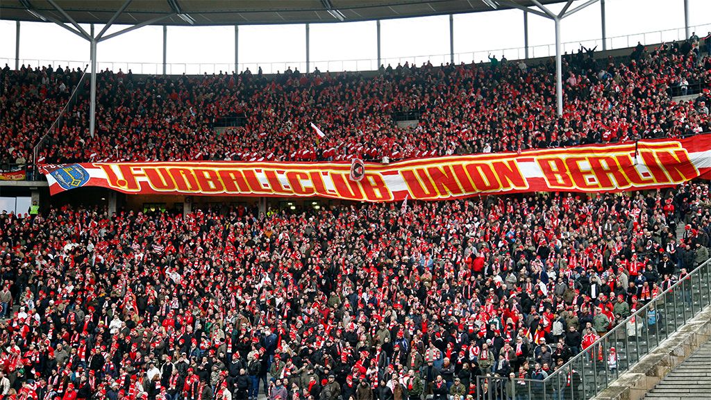 Fans des 1. FC Union Berlin stehen in einem Stadion und halten ein großes Banner hoch.