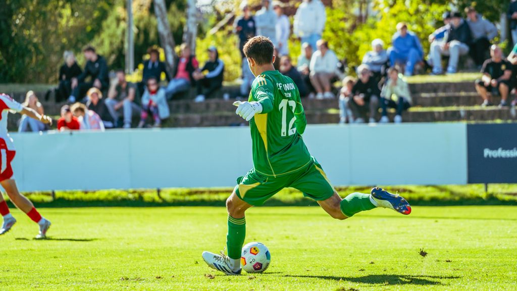 Ein Torwart in grüner Spielkleidung tritt den Ball auf einem Fußballfeld, während Zuschauer im Hintergrund stehen.
