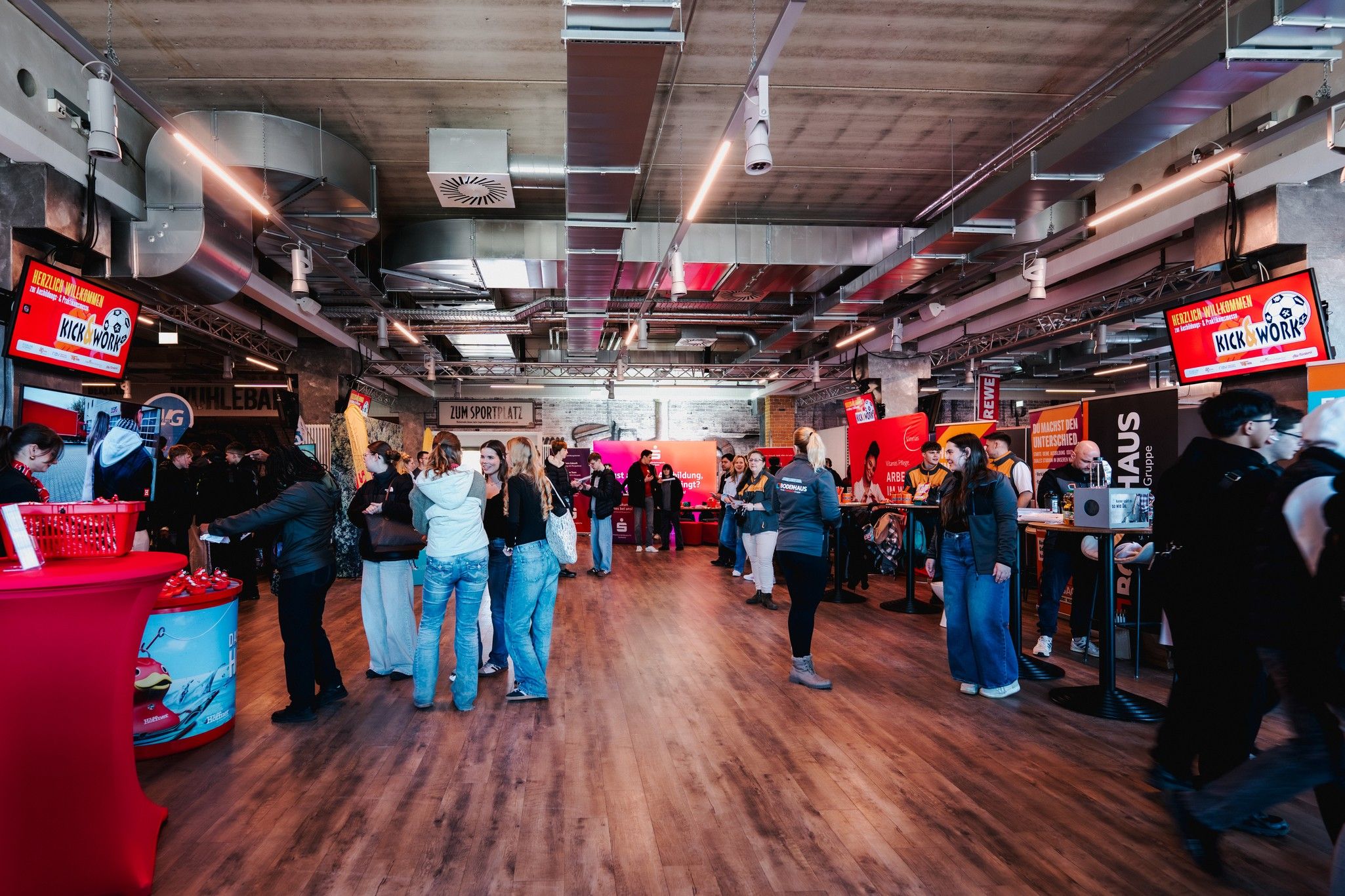 Menschen in einem modernen Veranstaltungsraum bei einer Jobmesse, umgeben von Ständen und Bildschirmen.