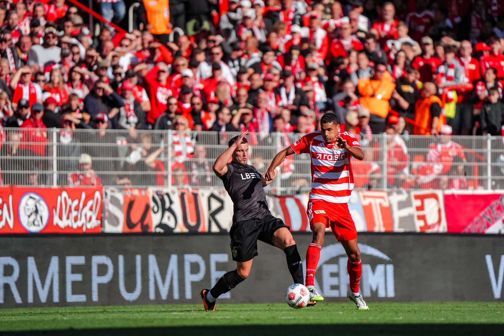 Spielszene im Fußball: Ein Spieler in schwarzem Trikot beim Zweikampf mit einem Spieler in rot-gestreiftem Trikot vor jubelnden Fans.