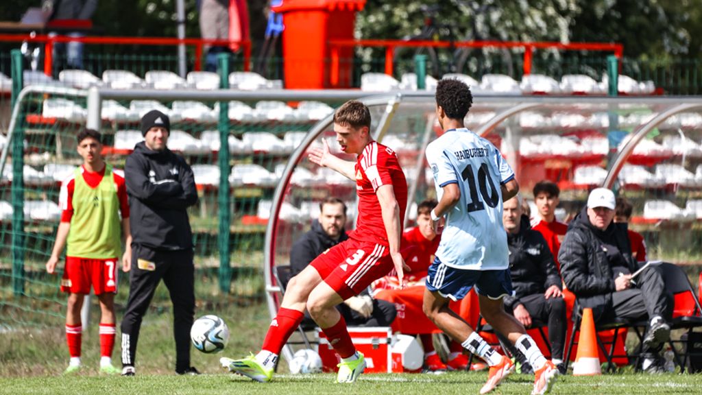 Ein Fußballspieler in rotem Trikot dribbelt den Ball, während ein Gegner in blauem Trikot ihn verfolgt. Zuschauer und Trainer sind im Hintergrund.