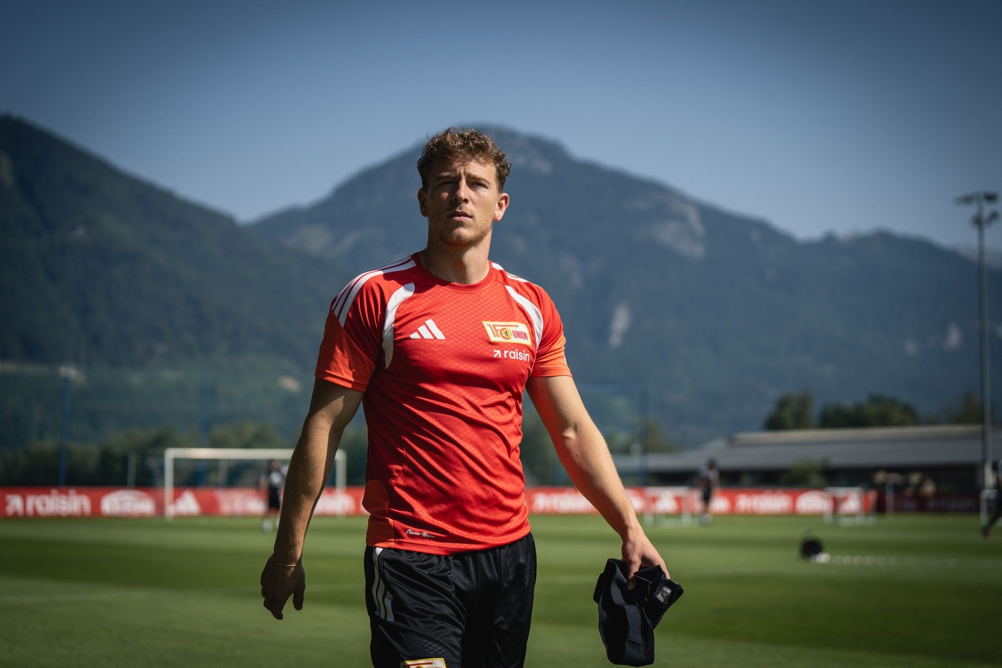 Ein junger Fußballspieler in rotem Trikot geht auf einem Trainingsplatz mit Bergen im Hintergrund.