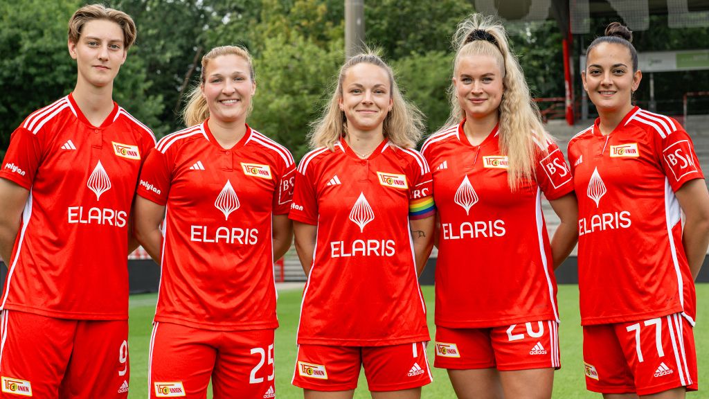 Fünf Fußballspielerinnen in roten Trikots mit dem Logo von Union Berlin stehen lächelnd auf einem Stadionfeld.