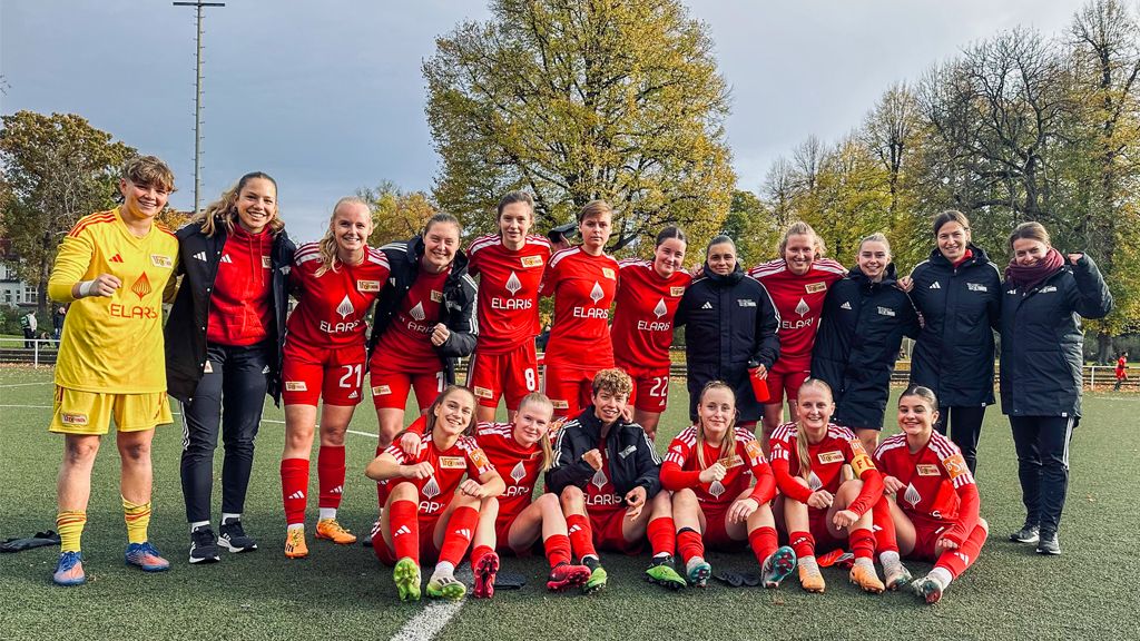 Eine Frauenfußballmannschaft in roten Trikots posiert lächelnd auf einem Sportplatz, einige knien, andere stehen.
