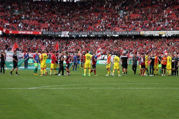 Fußballmannschaften stehen auf dem Spielfeld. Fans jubeln im Hintergrund, während Spieler sich die Hand geben.