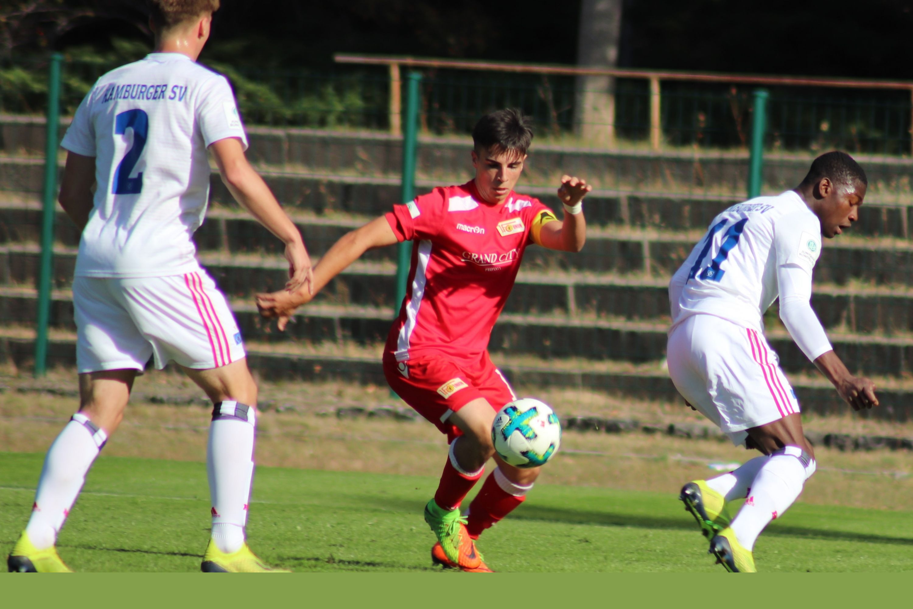 Zwei Fußballspieler in weißen Trikots verfolgen einen Spieler in rotem Trikot auf einem grünen Sportplatz.