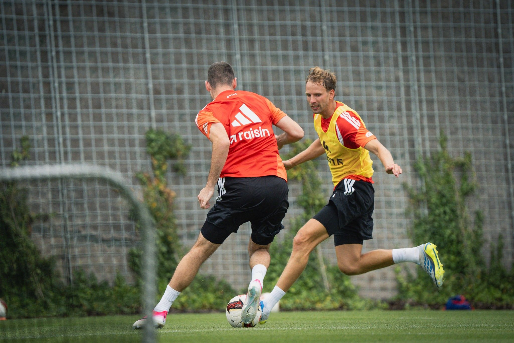 Zwei Fußballspieler in Training, einer dribbelt den Ball, während der andere ihn verfolgt. Hintergrund mit Tor und grüner Wiese.