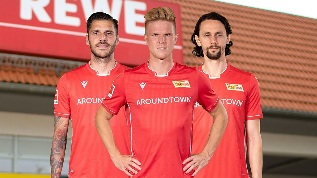 Drei Fußballspieler in roten Trikots stehen vor einem REWE-Supermarkt.