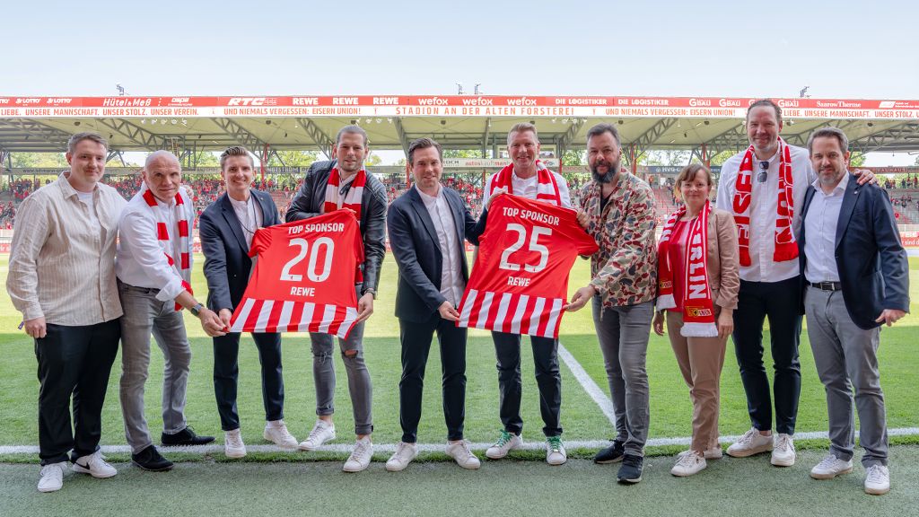 Zehn Personen stehen auf einem Fußballfeld, halten Trikots mit den Nummern 20 und 25 und tragen rote Schals. Umfeld eines Stadions sichtbar.