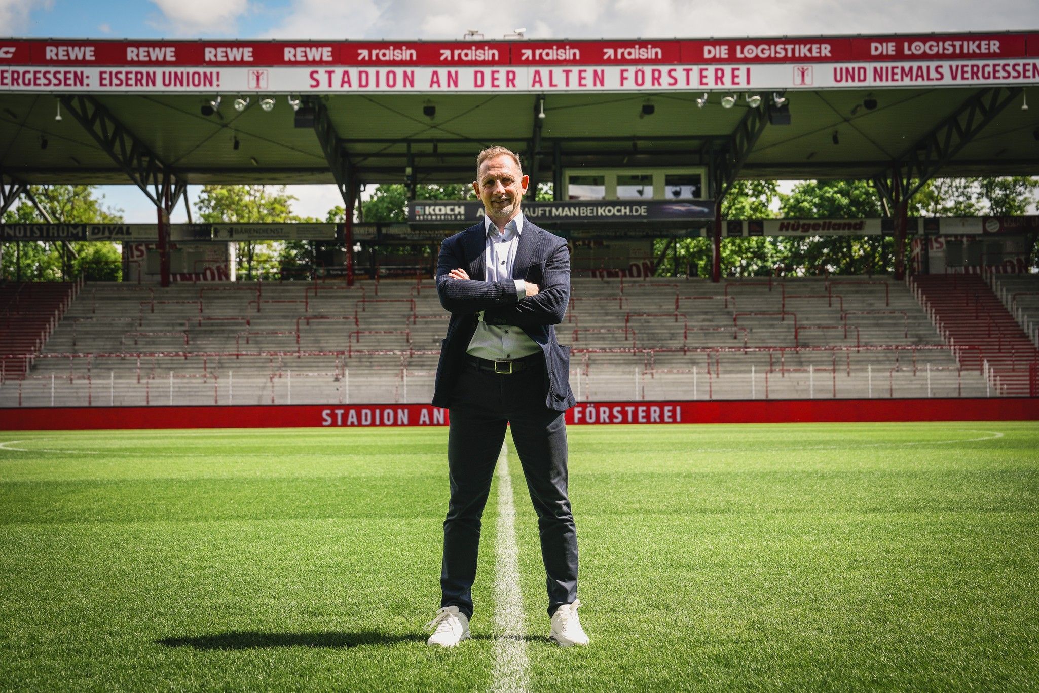 Mann steht lächelnd auf einem Fußballfeld im Stadion, umgeben von leeren Tribünen und Werbebannern.