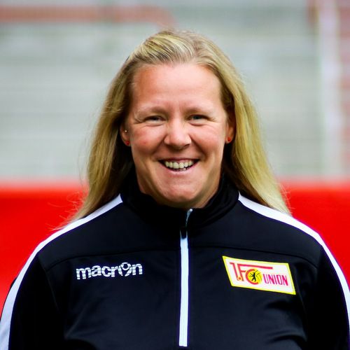 Eine lächelnde Frau mit blonden Haaren trägt eine schwarze Trainingsjacke mit einem Logo auf der Brust.