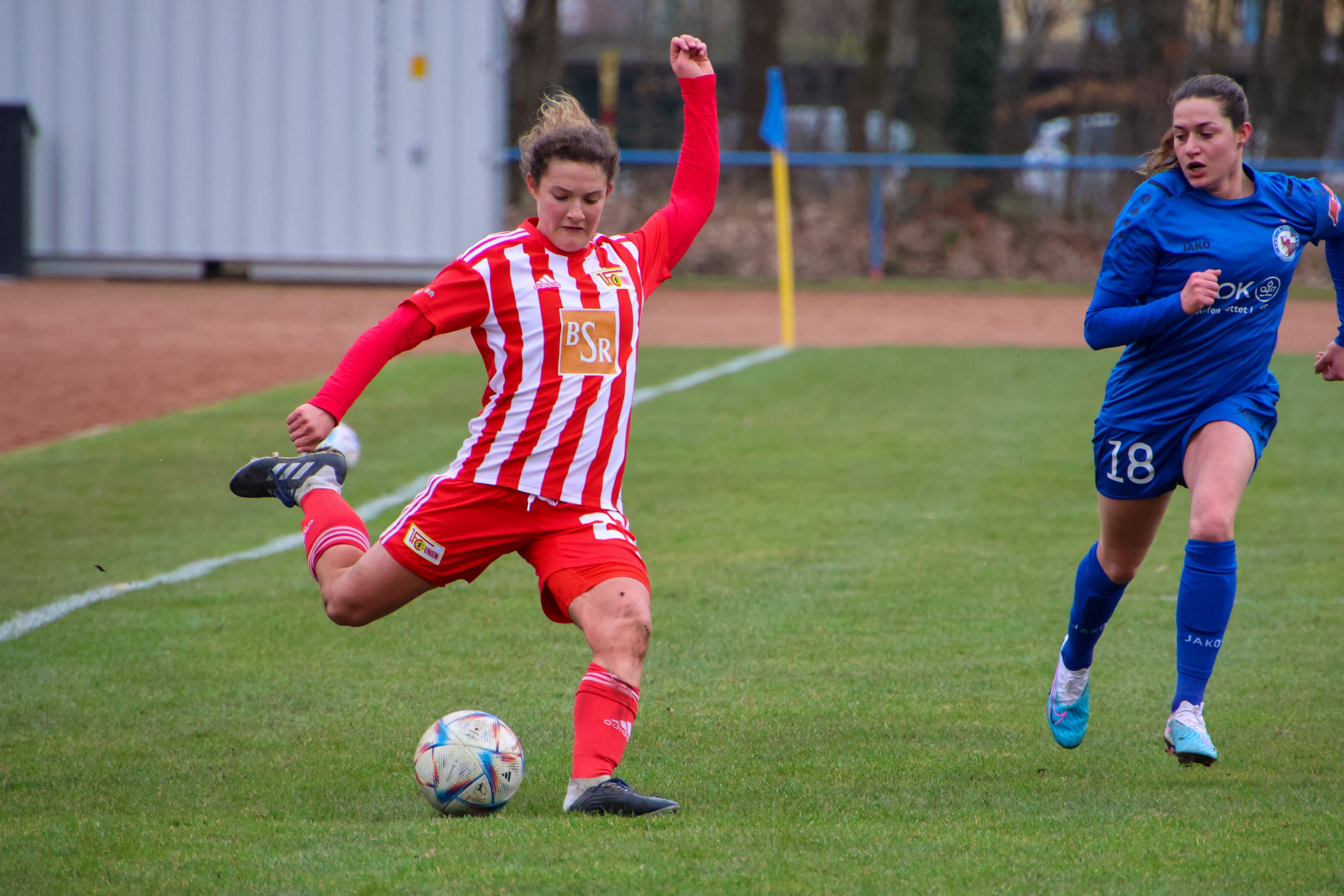 Eine Fußballspielerin in rot-weißer Uniform tritt den Ball, während eine andere Spielerin in blauer Uniform sich dem Ball nähert.