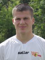 Mann mit kurzen, hellen Haaren, der in einem weißen Sporttrikot vor grünem Hintergrund lächelt.