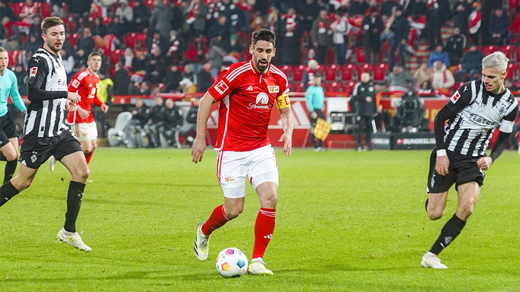 Ein Fußballspieler in rotem Trikot dribbelt den Ball, während zwei gegnerische Spieler in schwarz-weißen Trikots ihm folgen.