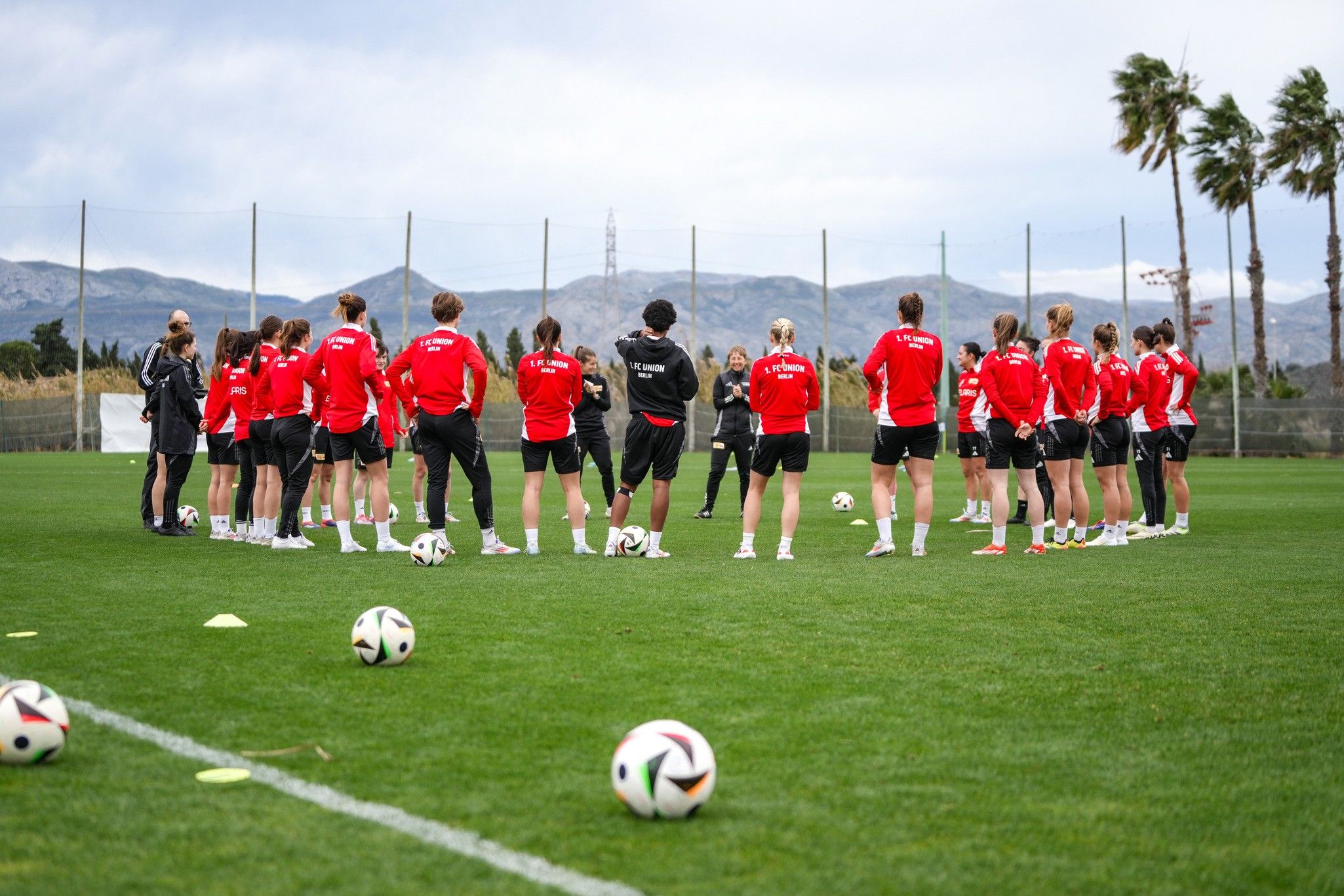 Trainingssitzung einer Frauensoccer-Mannschaft auf dem Platz mit mehreren Spielerinnen in roten Trikots und Bällen auf dem Boden.
