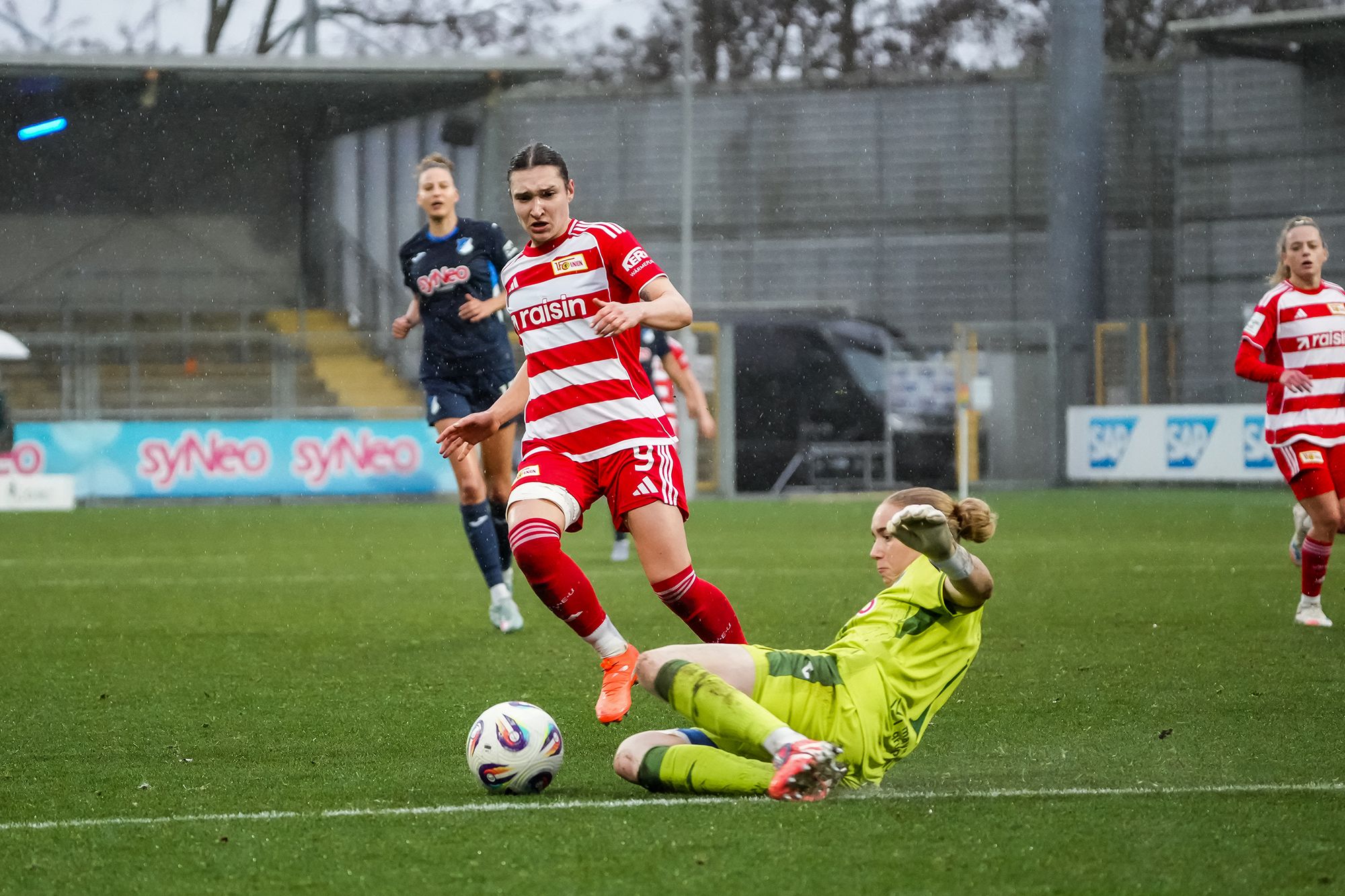 Eine Spielerin in rot-weißer Uniform greift in einem Fußballspiel an, während die Torhüterin im grünen Trikot einen Schuss abwehrt.