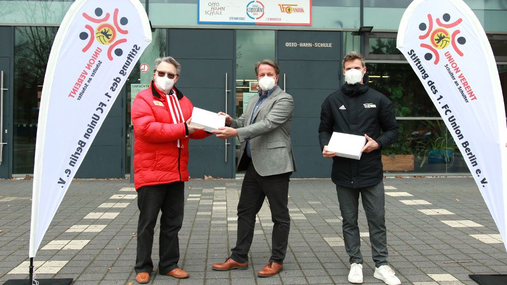 Drei Männer in Masken stehen vor einem Gebäude, übergeben sich Geschenke, umgeben von Fahnen mit Vereinslogos.