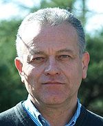 Ein Mann mit grauen Haaren, neutralem Gesichtsausdruck, trägt einen dunklen Pullover und eine hellblaue Hemdbluse, vor unscharfem grünem Hintergrund.