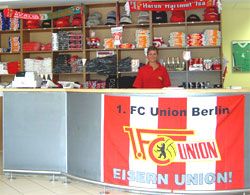 Ein Verkaufsstand mit einem roten Tisch und einem Banner des 1. FC Union Berlin. Eine Person steht hinter dem Tisch.