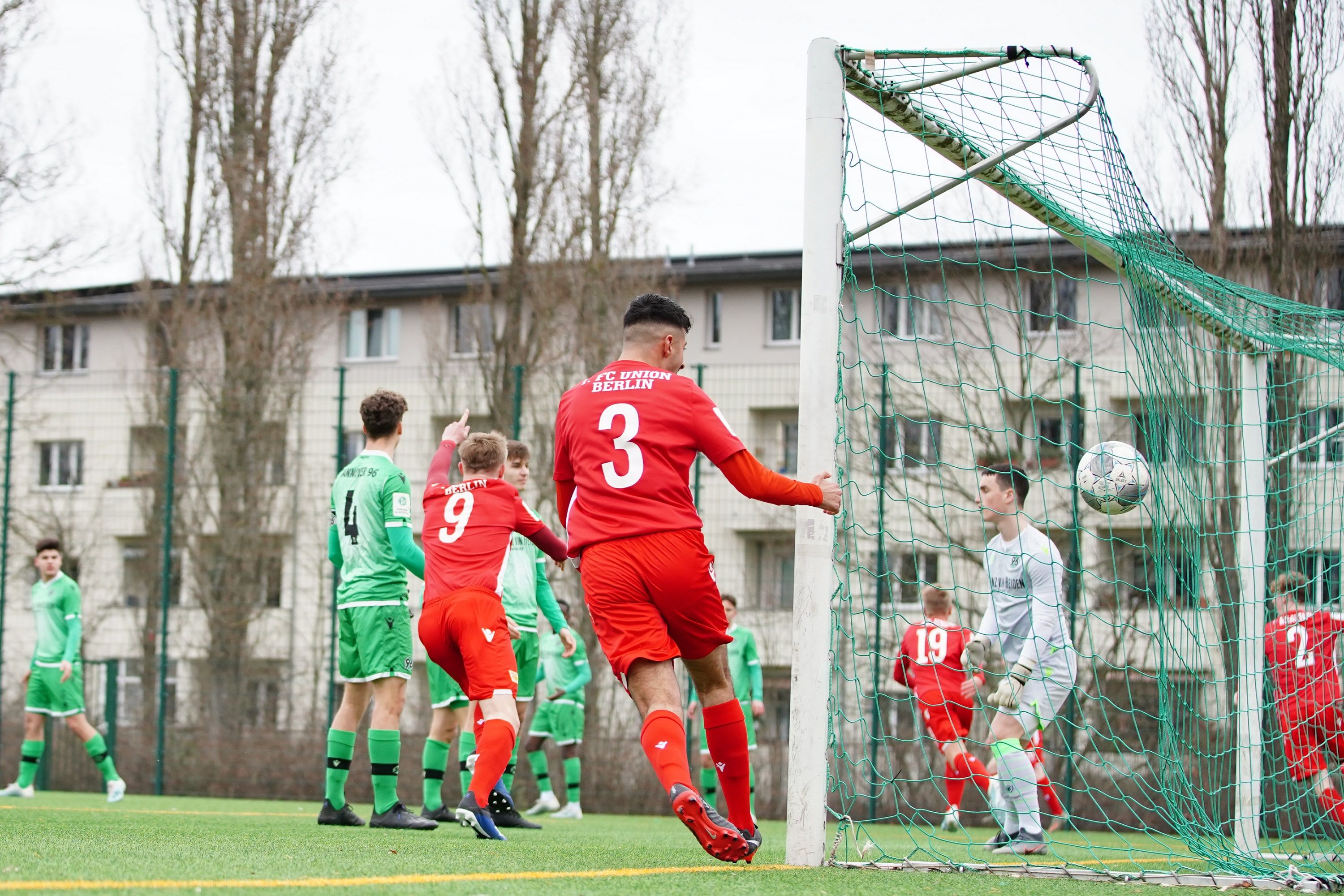 Ein Spieler in rotem Trikot erzielt ein Tor, während andere Spieler in grünen Trikots zuschauen.