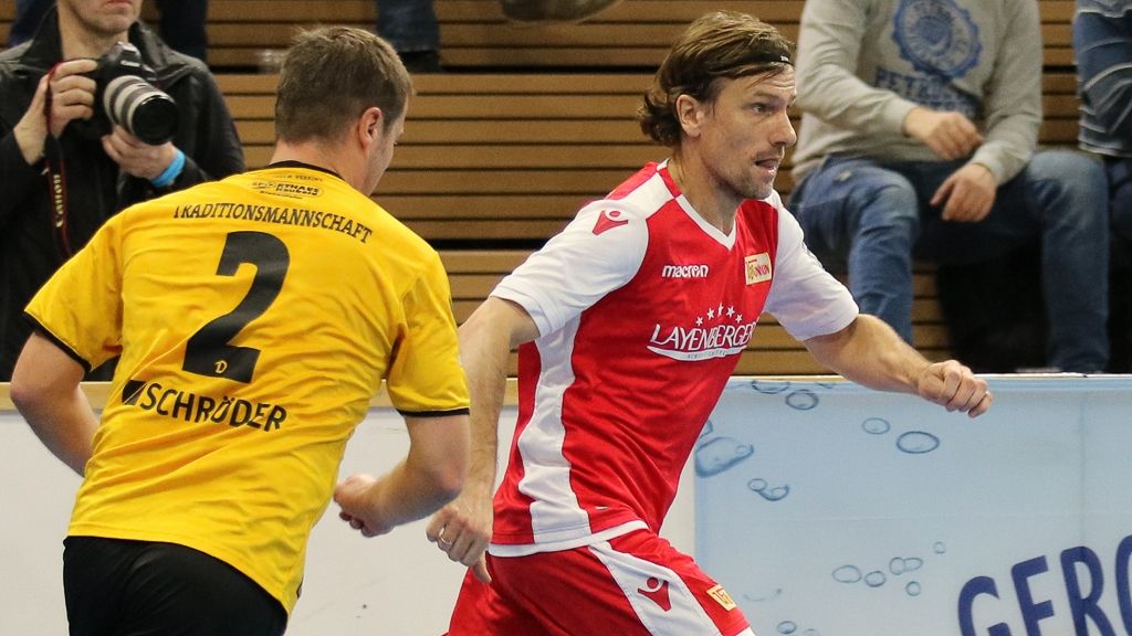 Zwei Handballspieler im Wettkampf; einer in gelbem Trikot, der andere in rotem Trikot. Hintergrund mit Zuschauern.