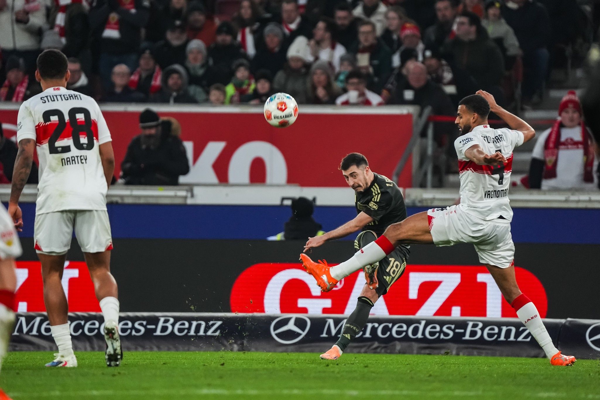 Spielszene: Ein Spieler schießt den Ball, während ein Gegenspieler versucht, ihn zu blocken, im Hintergrund jubelnde Fans.