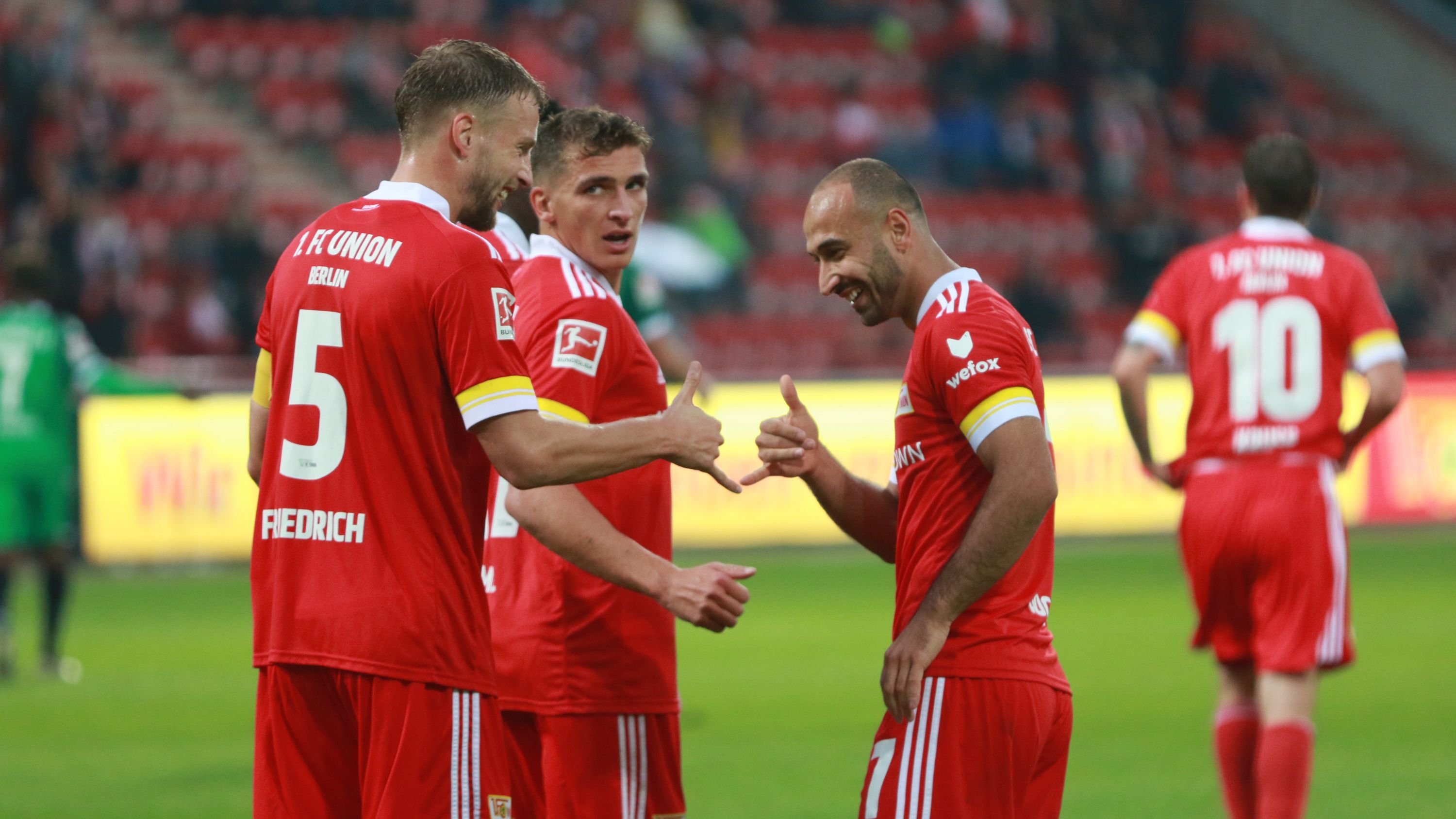 Drei Fußballspieler in roten Trikots stehen auf dem Spielfeld und zeigen sich gegenseitig das ‚Daumen hoch‘-Zeichen.