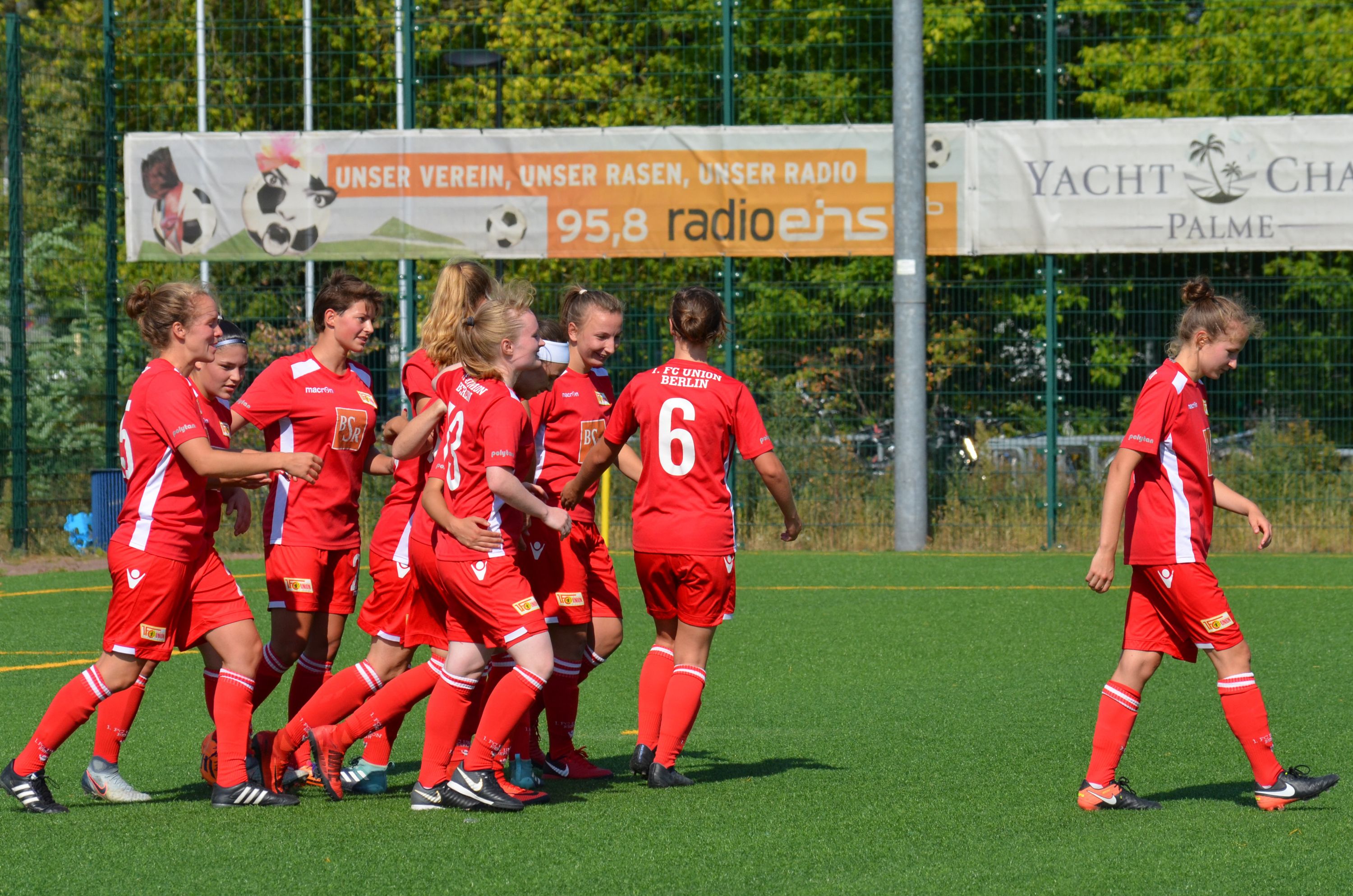 Frauenfußballmannschaft in roten Trikots feiert einen Torerfolg auf einem Kunstrasenplatz.