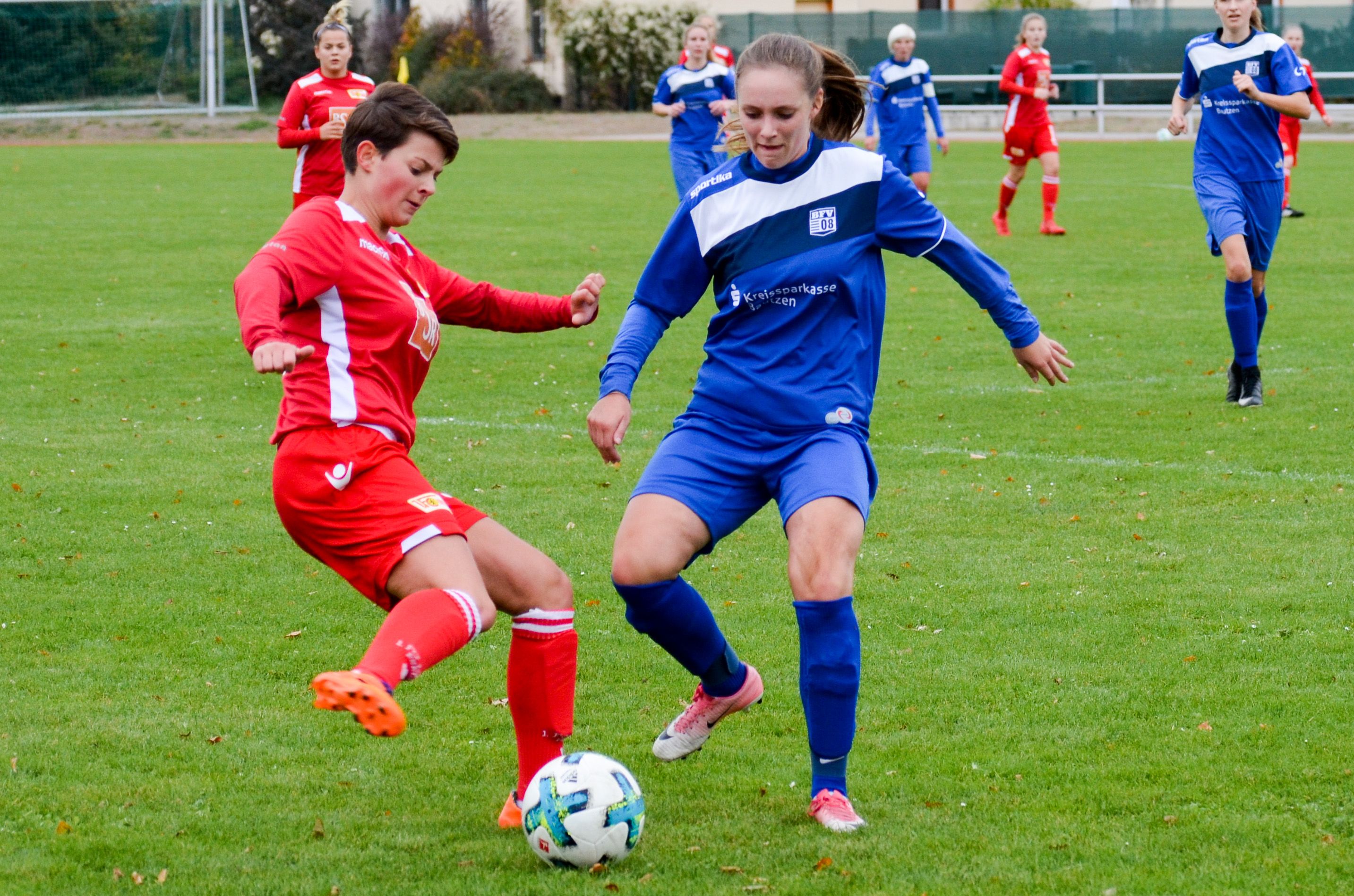 Zwei Frauen spielen Fußball. Sie kämpfen um den Ball. Sie stehen auf einem grünen Platz. Sie tragen unterschiedliche Trikots.