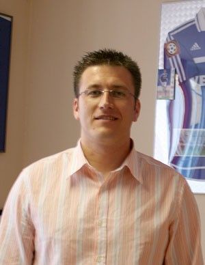 Ein Mann mit Brille und gestreifter Hemd steht vor einem Wandbild mit Fußballtrikots.