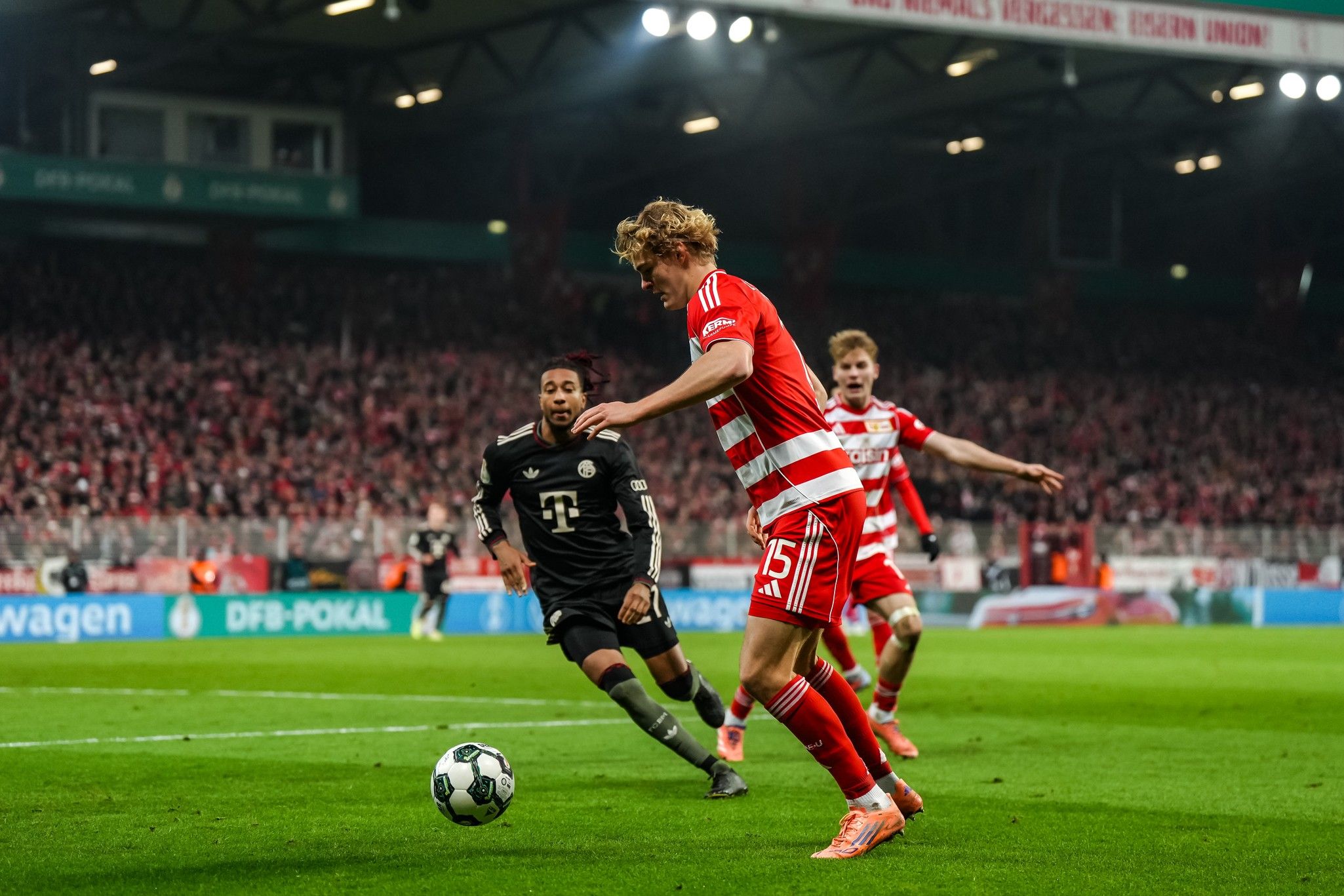 Spielszene im Fußball: Ein Spieler in roter und weißer Uniform dribbelt, während ein Gegner in schwarzer Uniform ihn verfolgt.