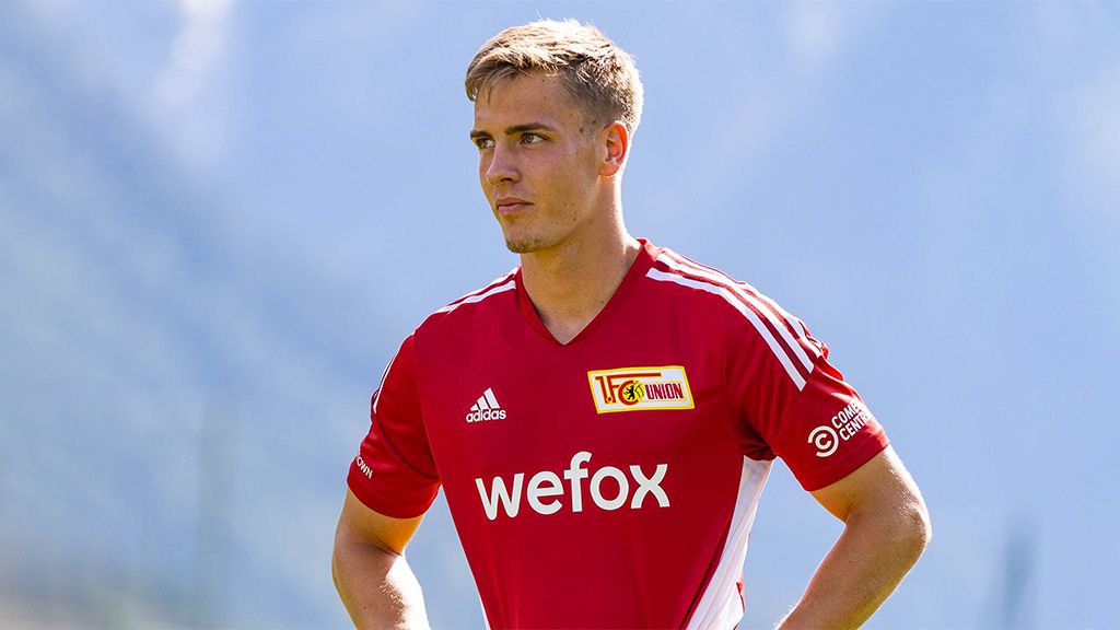 A young man in a red football jersey stands thoughtfully outdoors with mountains in the background.