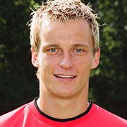 Ein junger Mann mit blonden, kurz geschnittenen Haaren, lächelt und trägt ein rotes Sportshirt vor einem grünen Hintergrund.