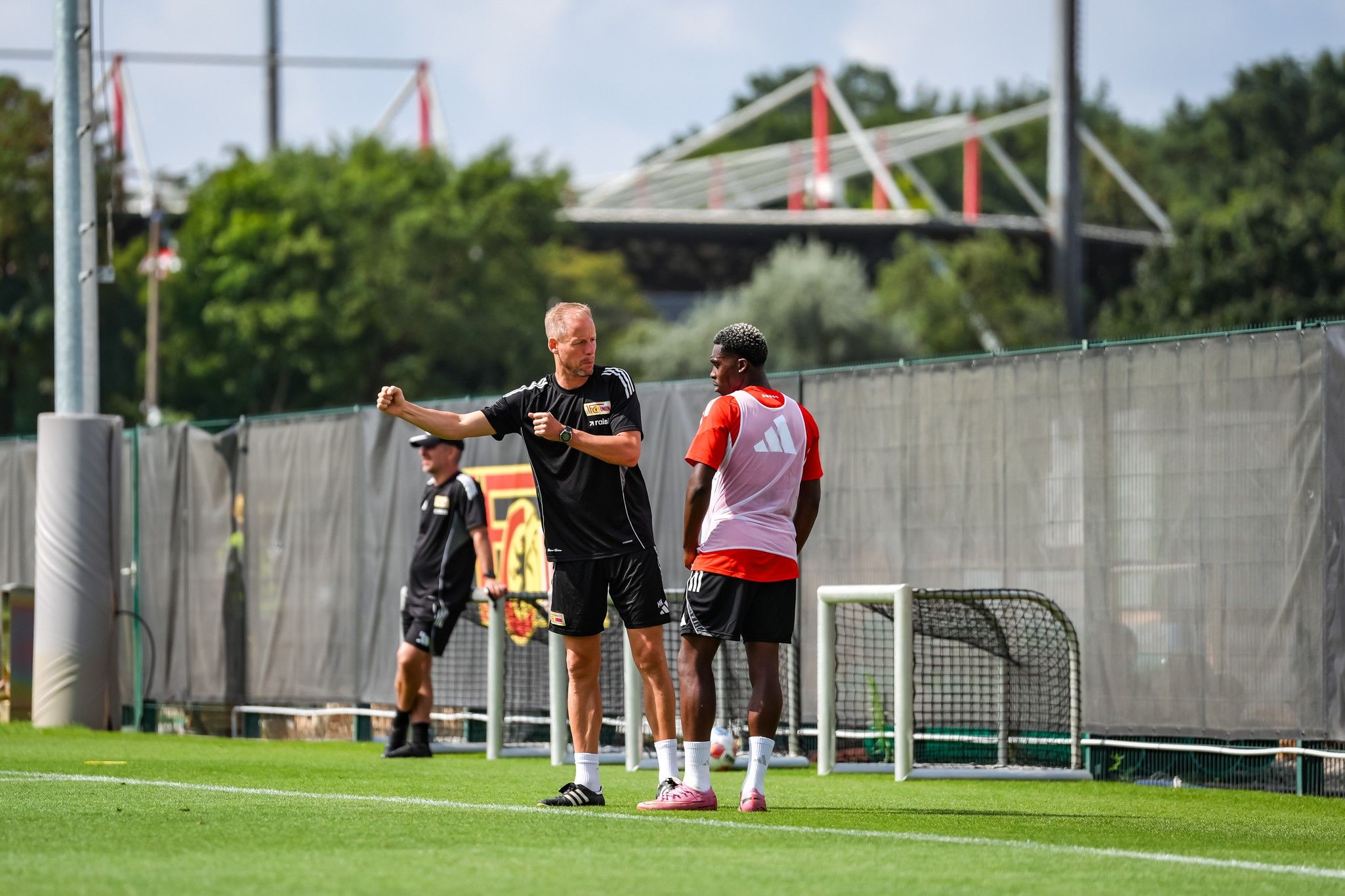 Trainer spricht mit einem Spieler auf dem Trainingsplatz, im Hintergrund sind die Umrisse eines Stadions sichtbar.