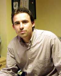 Ein Mann mit kurzen braunen Haaren sitzt in einem Büro und schaut in die Kamera. Er trägt ein helles Hemd.