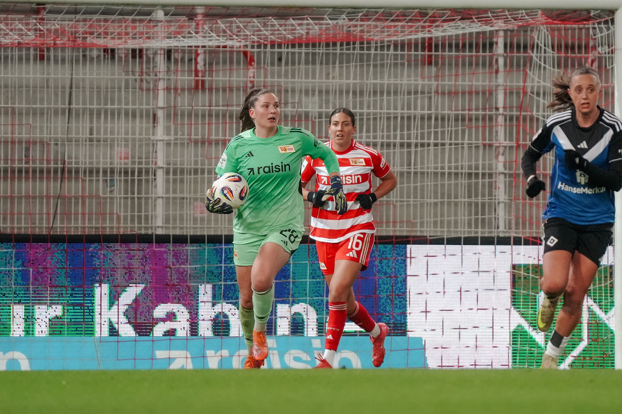 Eine Torhüterin in grüner Kleidung hebt den Ball, während zwei Spielerinnen des anderen Teams hinter ihr herlaufen.