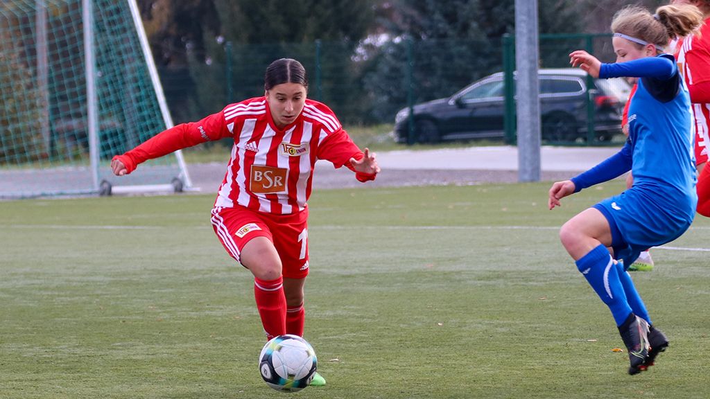 Eine Fußballspielerin in rot-weißem Trikot dribbelt den Ball auf einem Sportplatz, während eine Gegnerin in blauem Trikot angreift.
